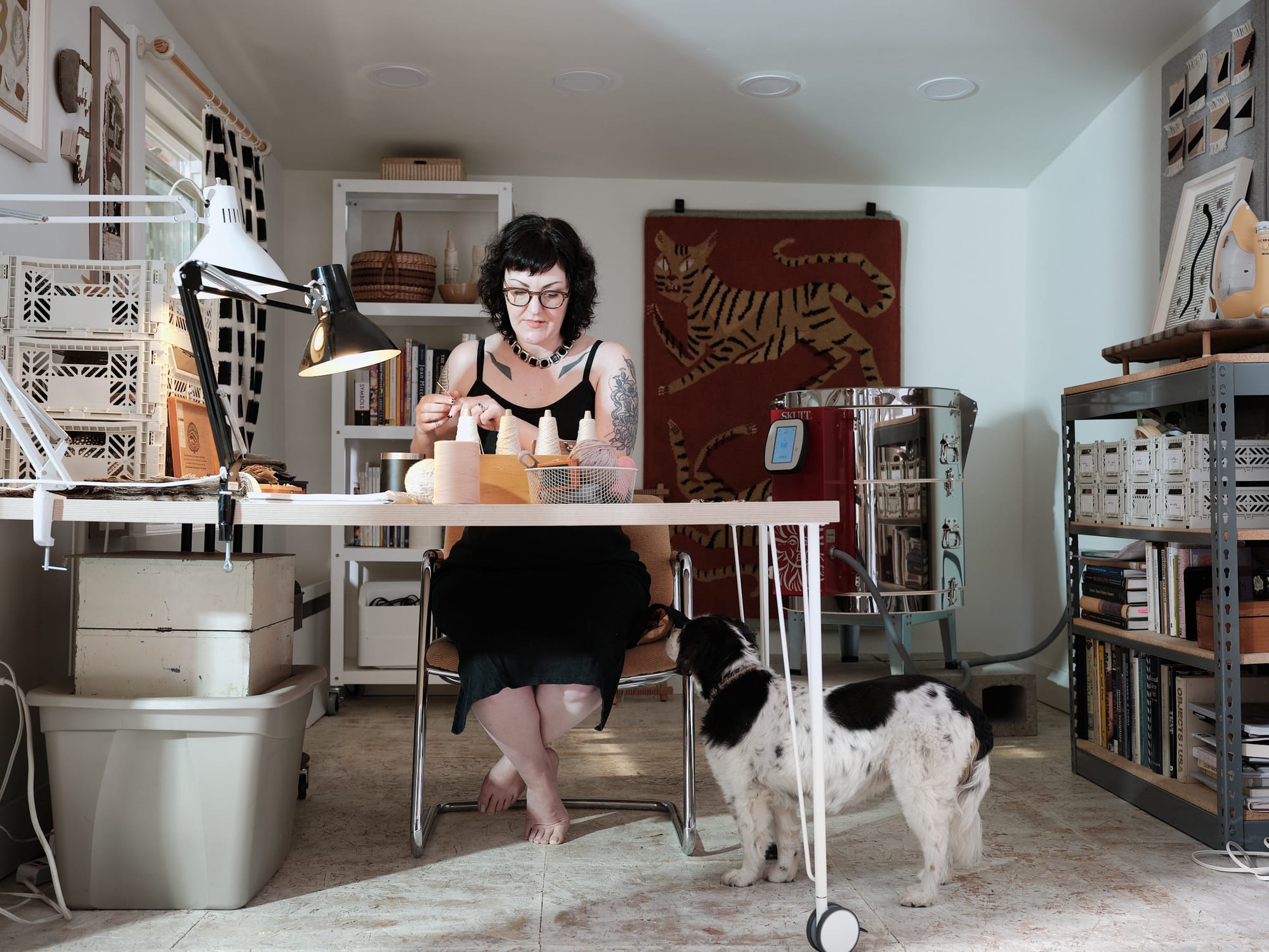 A tattooed woman with dark hair and glasses works at a craft table in an art studio, surrounded by spools, books, and artwork. A black-and-white dog stands at her feet.