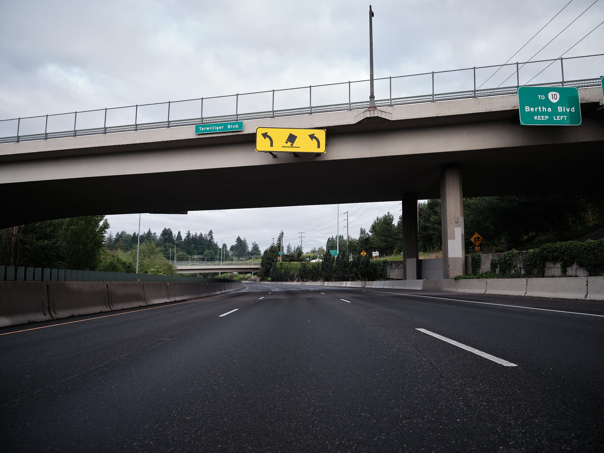 A completely empty freeway passes beneath the Terwilliger Boulevard overpass in Portland, Oregon. A yellow warning sign depicting a tipping truck flanked by turn arrows hangs from the bridge deck. A green highway sign on the right reads "To Route 10, Bertha Blvd, Keep Left." Evergreen trees and an overcast sky are visible in the distance.