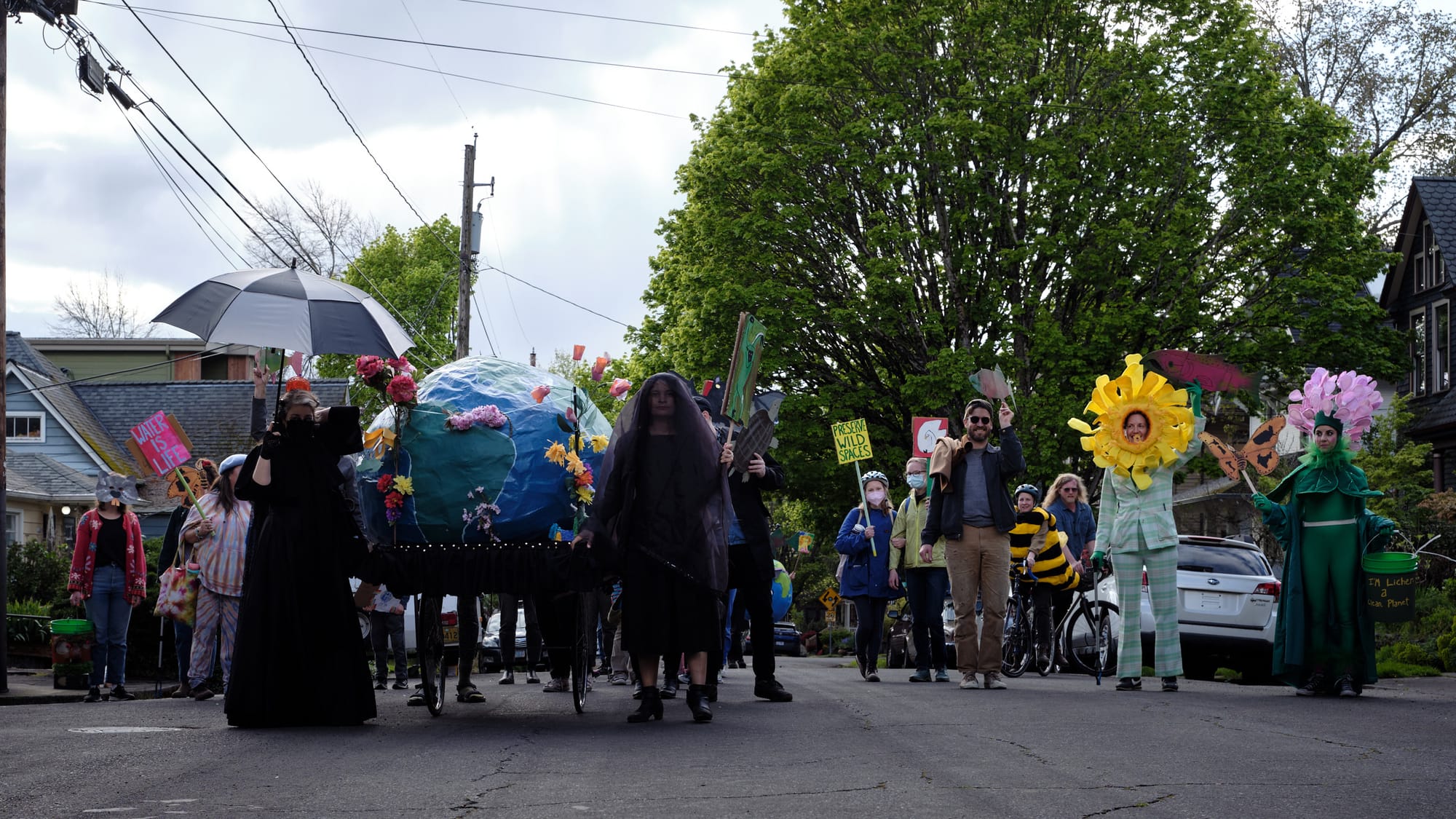 A group of environmental activists dressed in black participate in a community parade in Portland, Oregon, carrying a large Earth globe decorated with flowers on a wheeled cart while walking down a residential street lined with trees and houses.
