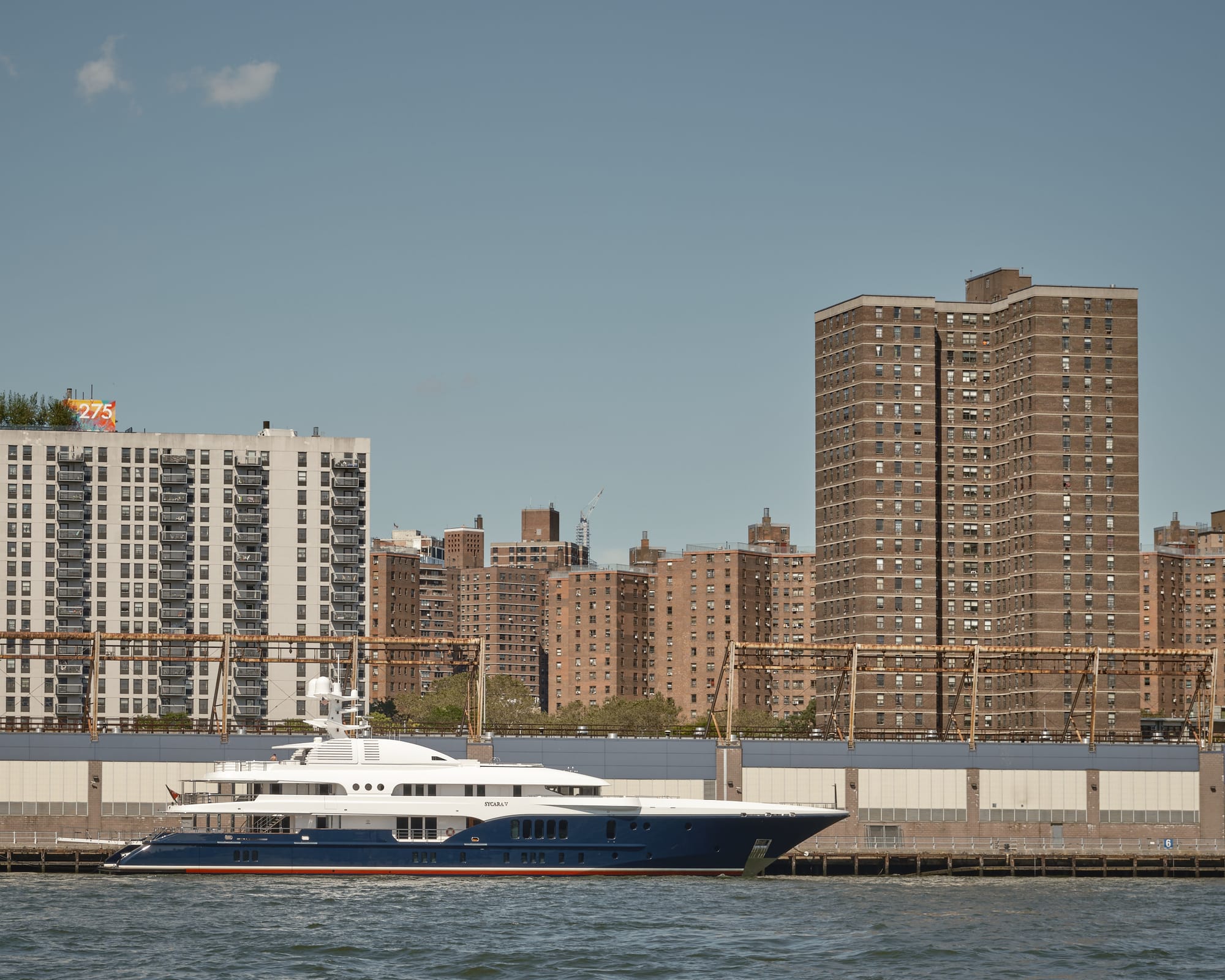 A large luxury superyacht with a deep navy blue hull, red waterline stripe, and white superstructure is moored at a pier along the Manhattan waterfront, photographed from across the East River in Brooklyn. The vessel's name appears to read "Sycara V" on the hull. Behind the yacht, rusted industrial pier infrastructure and a large grey waterfront building line the shore. Rising above are multiple massive brown brick mid-rise residential housing towers characteristic of Lower East Side public housing complexes. A colorful rooftop mural bearing the number "275" is visible on one of the shorter buildings at left. The sky is clear and hazy blue, with a single small cloud. The contrast between the opulent private yacht and the dense public housing towers behind it is stark.