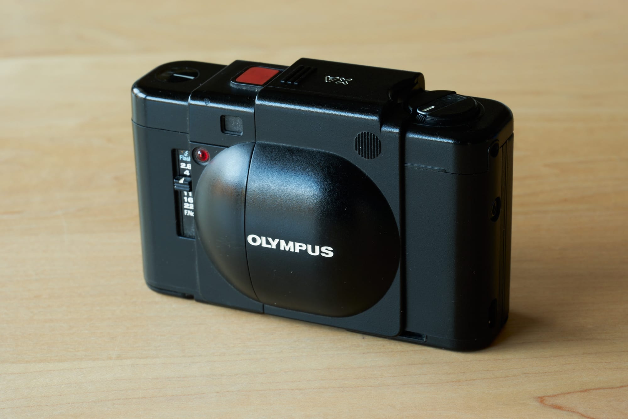 A pristine Olympus XA rangefinder camera rests on warm blonde wood, its charcoal metallic body gleaming under soft natural light. The compact 35mm camera's distinctive sliding lens cover reveals the aperture priority f/2.8 lens, while the red shutter release button provides a subtle accent against the matte black top plate. The intimate composition captures the camera at a three-quarter angle, emphasizing its legendary portable design that made it a favorite among street photographers.