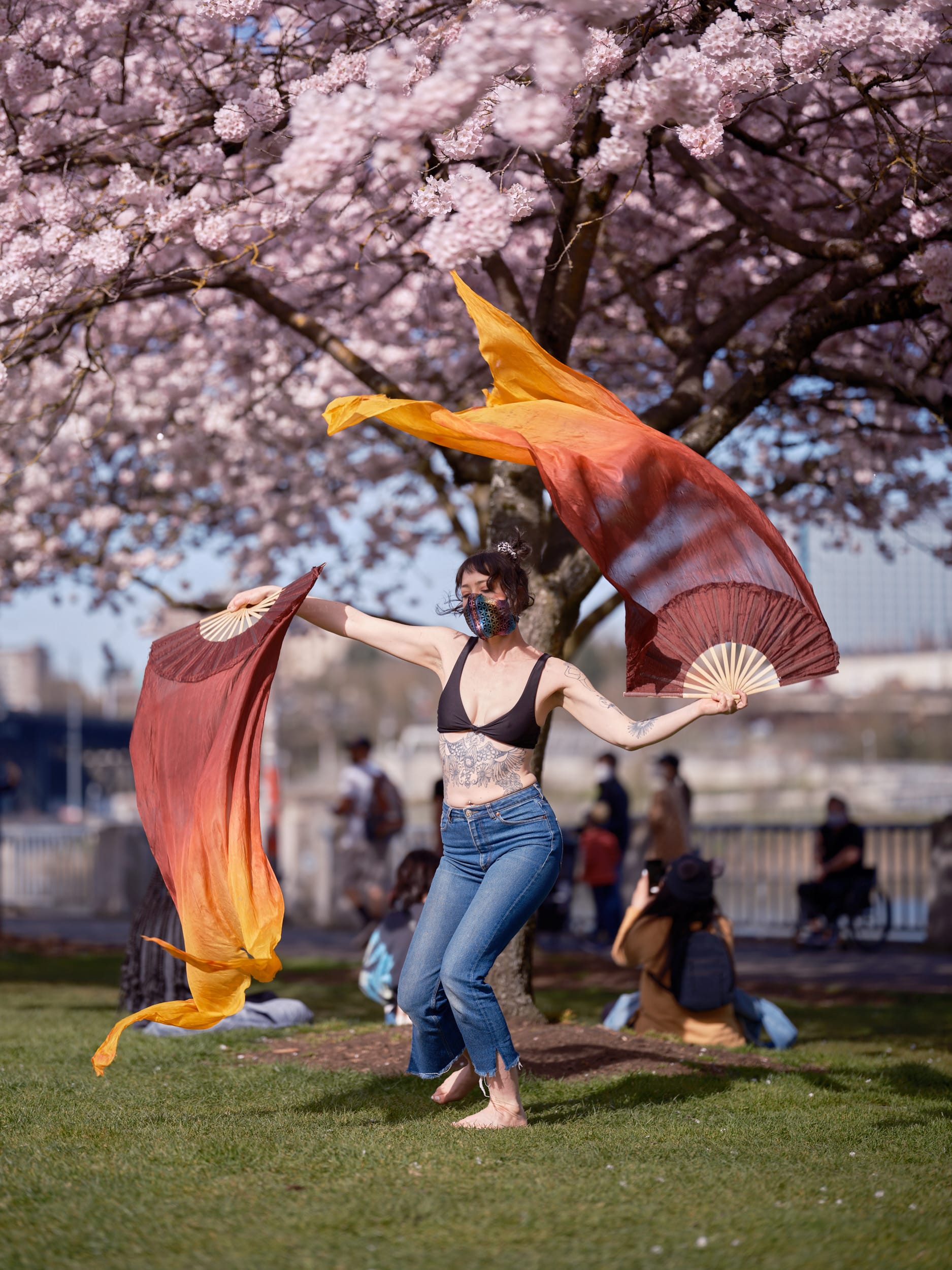 A masked performer moves gracefully through Tom McCall Waterfront Park, her flowing silk fans catching the golden light as they billow like flames against a canopy of delicate pink cherry blossoms. The dancer, wearing jeans and a dark top, creates a striking contrast between contemporary street style and traditional fan dance artistry. Behind her, the dreamy blur of spring visitors and the iconic waterfront setting frame this moment of cultural celebration in Portland's Old Town.