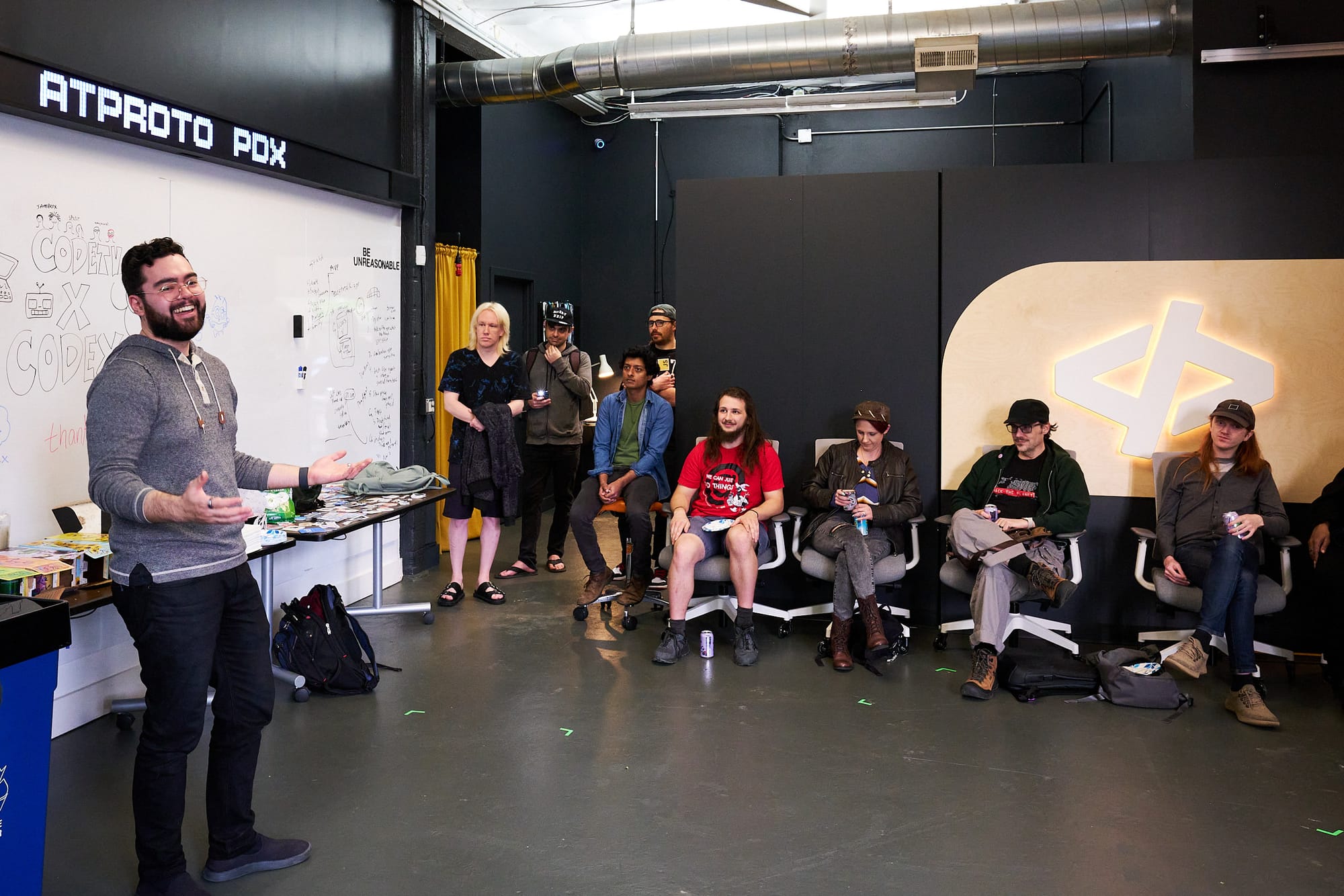 Graham stands in front of a whiteboard covered in Code TV doodles and the phrase "Be Unreasonable," gesturing as he welcomes the group. Attendees sit and stand in a loose semicircle listening, holding cans of drink. The ATPROTO PDX sign is visible at the top left.
