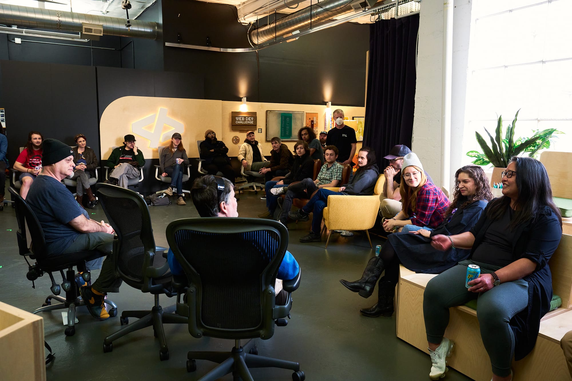 A wide view of roughly thirty people seated on Aeron chairs and low couches inside a television studio, facing toward a speaker off-frame. A large backlit "</>" Code TV logo is mounted on the back wall next to a "Web Dev Challenge" sign. Tall windows and a large snake plant on the right.