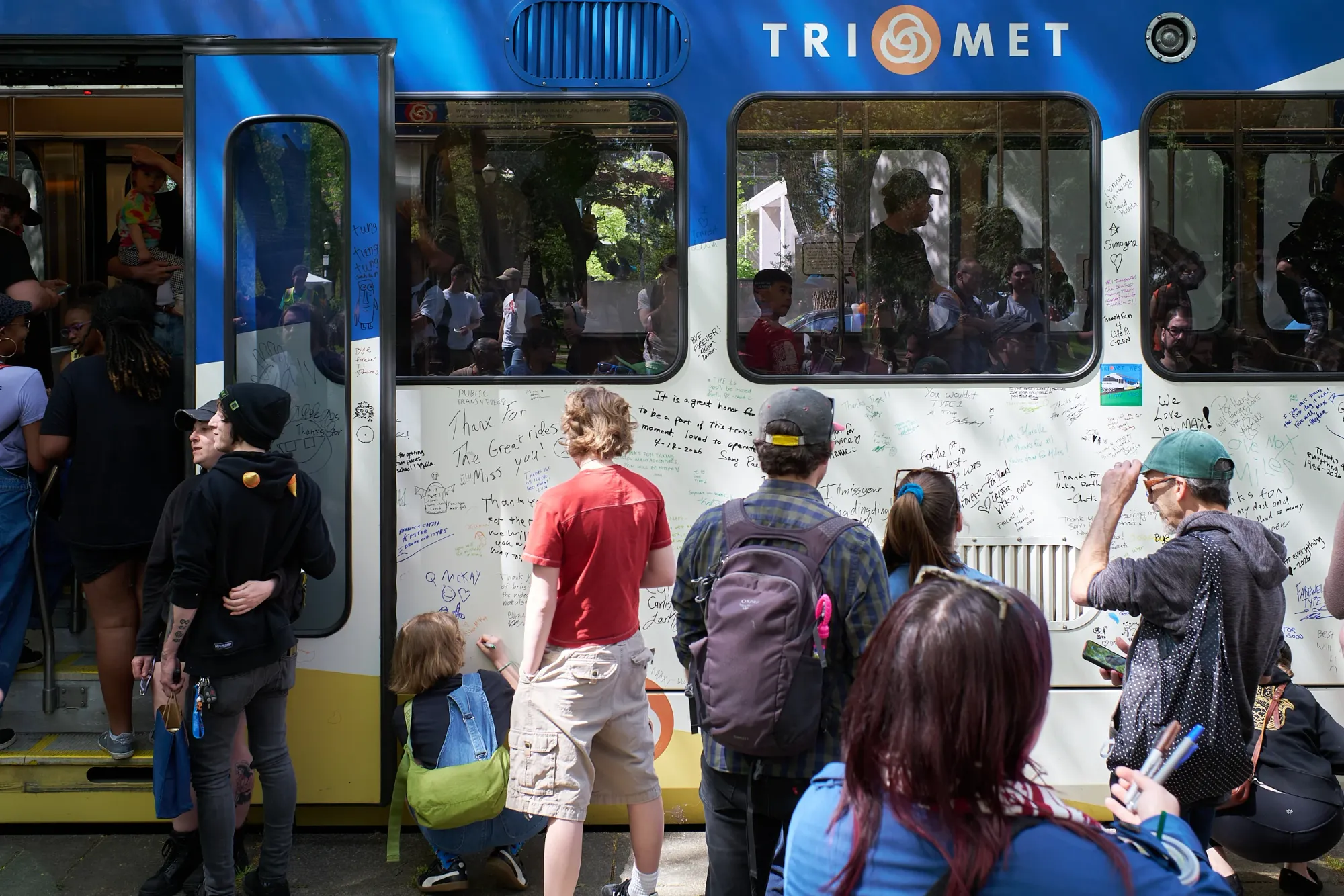 A crowd of Portlanders writes farewell messages in marker on the white side panels of TriMet MAX Type 1 car 124, which is nearly covered in handwritten signatures and tributes in many colors. A child kneels to sign near the bottom while adults stand writing and reading messages like "Thanx for the great rides" and "We love you, MAX." The train's open doors and TriMet logo are visible above the crowd on a sunny afternoon.