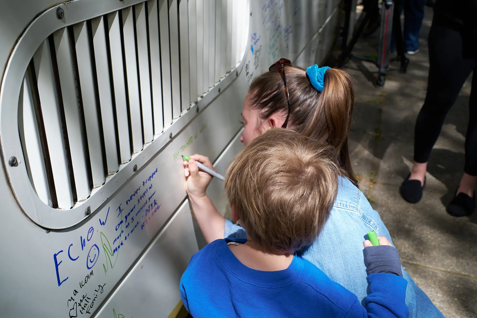 A woman in a denim jacket writes 'YOU TRAINED US WELL' in green marker on the side of MAX car 124 while her young child in a blue sweatshirt looks over her shoulder.