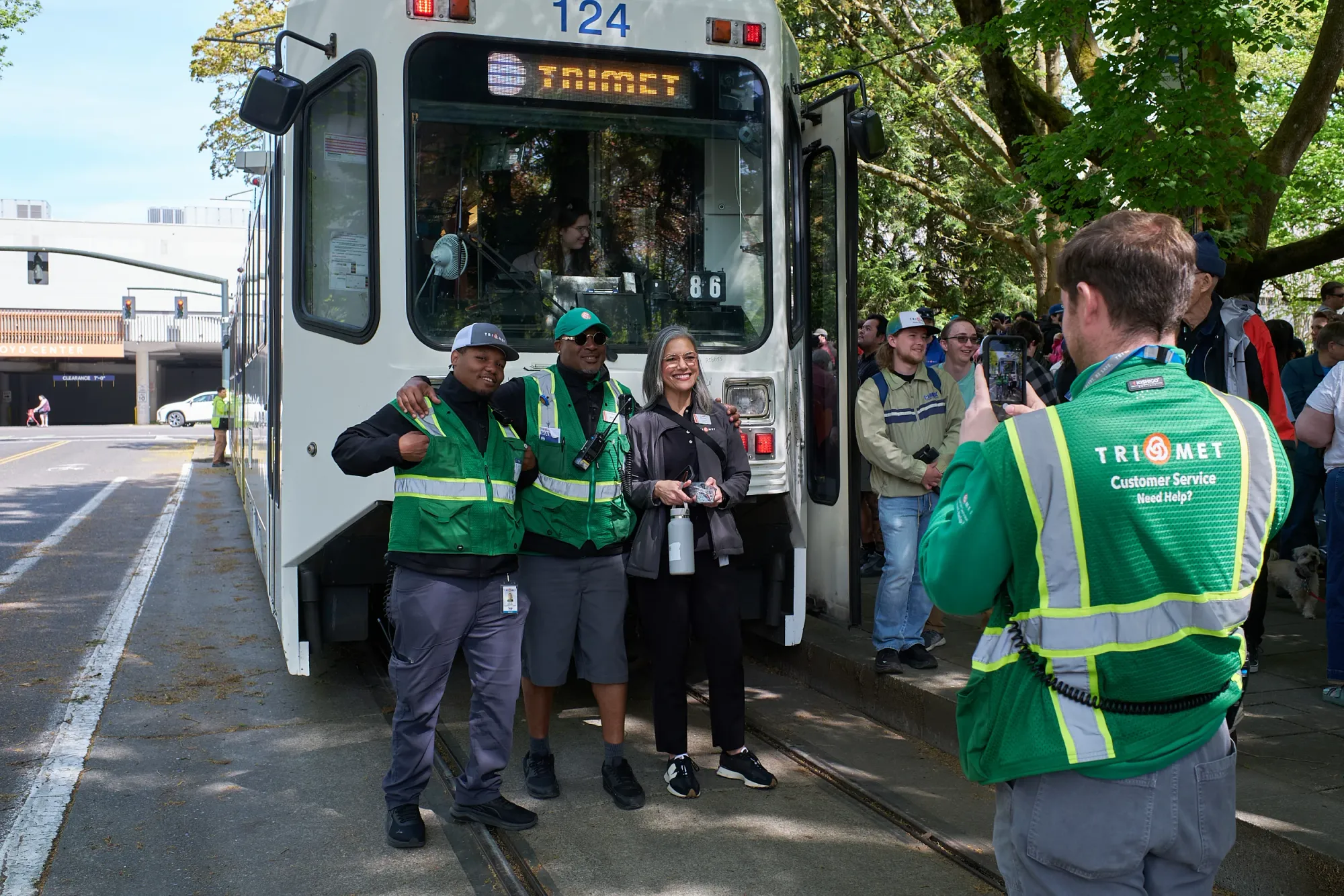 Three TriMet Customer Service representatives in green high-visibility vests stand with a supervisor in front of MAX Type 1 car 124 while a fourth representative takes their photo with a phone.