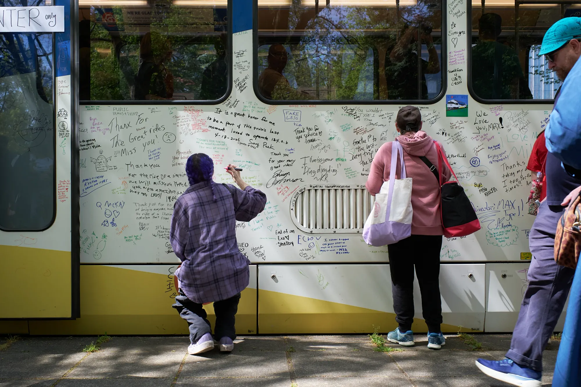 A person with a purple braid in a purple flannel kneels to sign MAX car 124 while another person in a pink hoodie stands beside them, both surrounded by densely written farewell messages and tributes in many colors of marker.