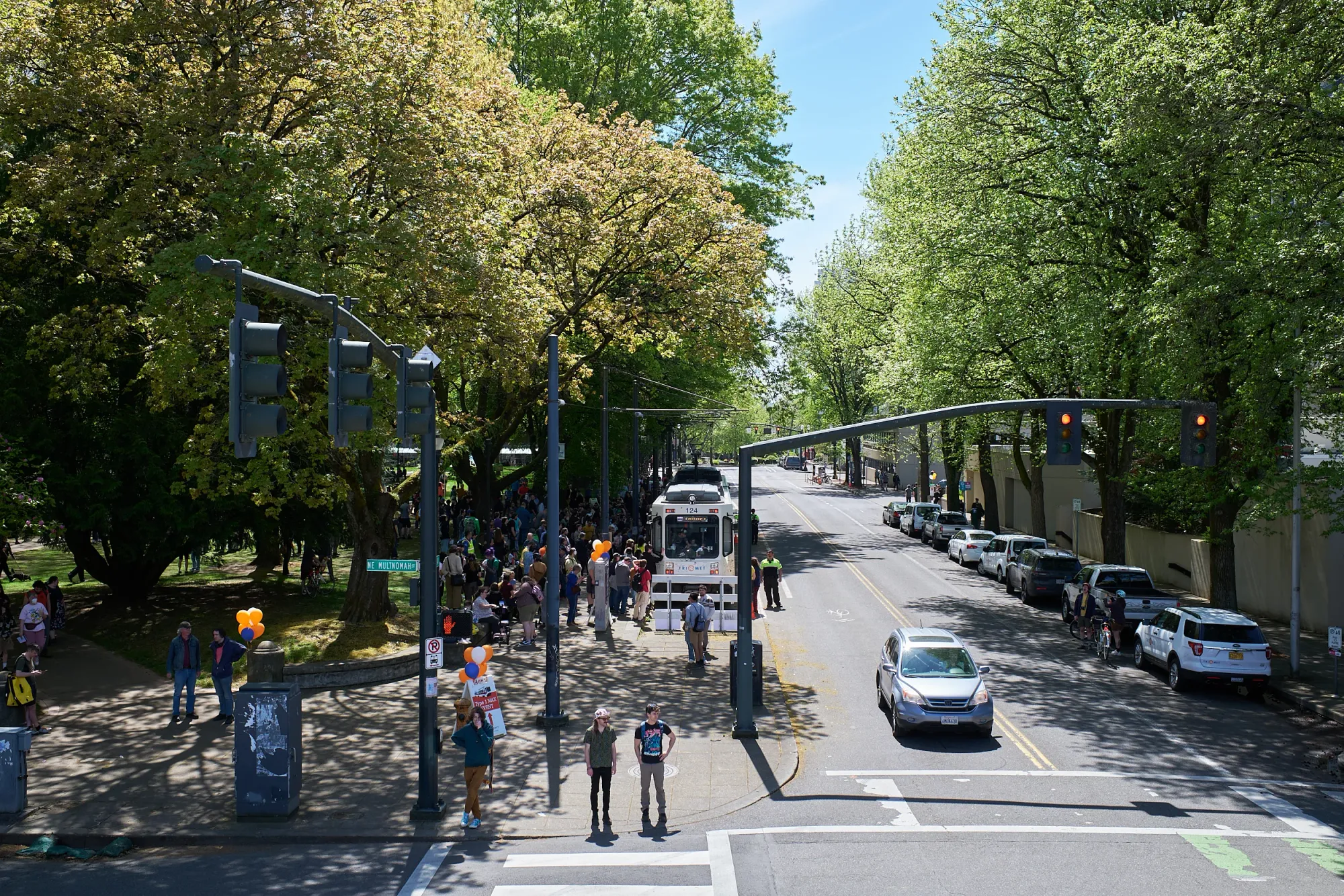 An elevated view down NE Multnomah Street on a sunny spring day, with MAX Type 1 car 124 stopped beneath a canopy of trees and a crowd gathered alongside it at Holladay Park.