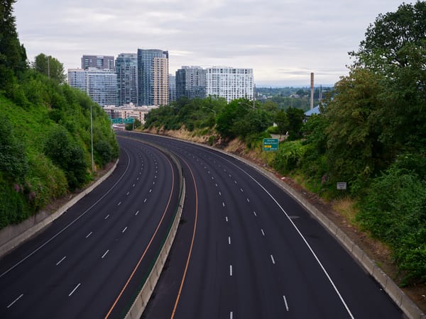 Empty lanes of I-5 curve through a green corridor in Portland, Oregon, with a South Waterfront exit sign and modern high-rise towers visible in the background.
