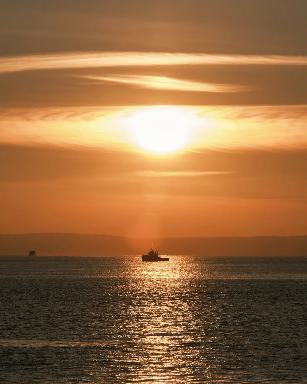 A tugboat is silhouetted against a vivid golden sunset over Elliott Bay in Seattle, Washington. The setting sun reflects in a shimmering path across the calm water. Wispy clouds stretch across the deep orange sky, and a faint landmass is visible on the distant horizon to the left.