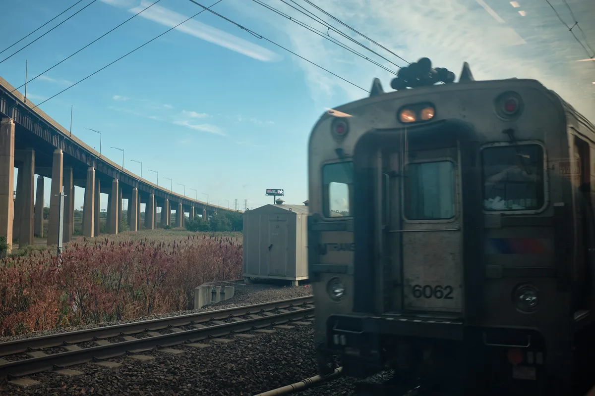 A close-up view of an NJ Transit commuter train car numbered 6062 passing on an adjacent track, photographed through a window near Kearny, New Jersey. A conductor is visible leaning against the glass inside the passing train. Wild reddish-pink vegetation lines the rail corridor in the foreground. A large elevated Interstate 95 viaduct stretches across the background on concrete columns under a blue sky with wispy clouds. A small utility shed and a distant billboard are visible between the train and the highway overpass.