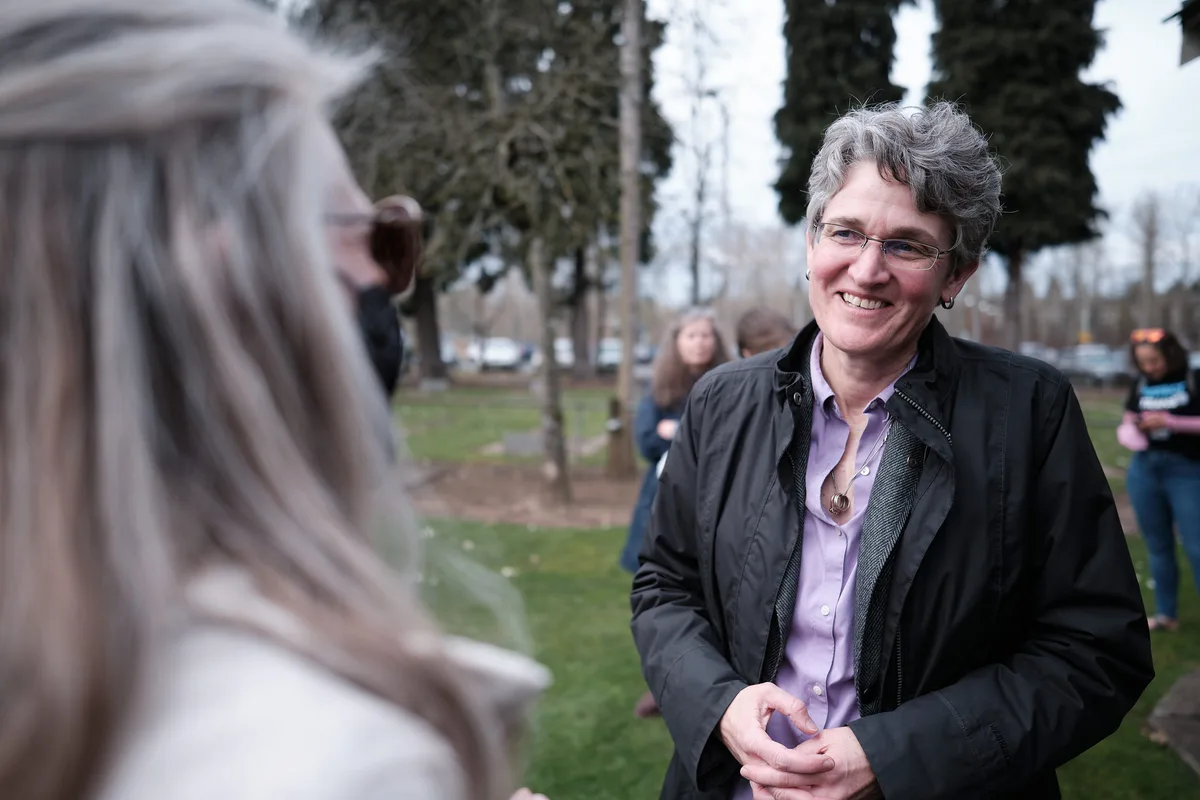 Congressional candidate Jamie McLeod-Skinner smiles warmly while speaking with a supporter at an outdoor campaign event. She has short grey hair, wire-rimmed glasses, and wears a black jacket over a lavender button-up shirt with a pendant necklace. The supporter is seen from behind in the blurred foreground. Other attendees and bare-branched trees are visible in the background.