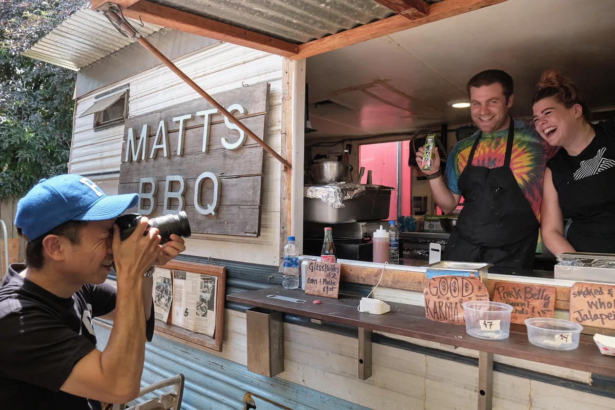 A Japanese tourist wearing a blue baseball cap photographs two laughing staff members at Matt's BBQ food cart on Mississippi Avenue in Portland, Oregon. The male staff member in a tie-dye shirt and black apron holds up a small Japanese snack item for the camera, while his female colleague beside him laughs. The rustic wood-sided cart displays a hand-lettered sign reading "Matt's BBQ," along with menu signs for pork belly and smoked jalapeño. Handwritten signs on the counter read "Good Karma" and "Glass Bottles $2."