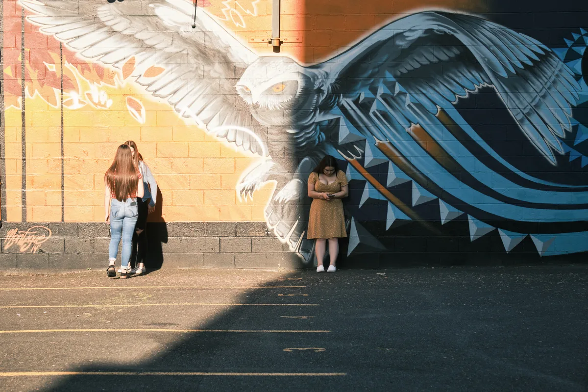 Against the Pearl District's vibrant street art, three figures maintain natural distances along a sun-dappled sidewalk, unknowingly embodying social distancing before the concept entered our collective consciousness. A magnificent owl mural with outstretched wings dominates the brick wall, its piercing gaze overseeing the scene as golden hour light bathes the urban canvas. Two women stand apart examining the artwork while a third figure retreats into shadow, creating an unintentional tableau of personal space and solitary contemplation.