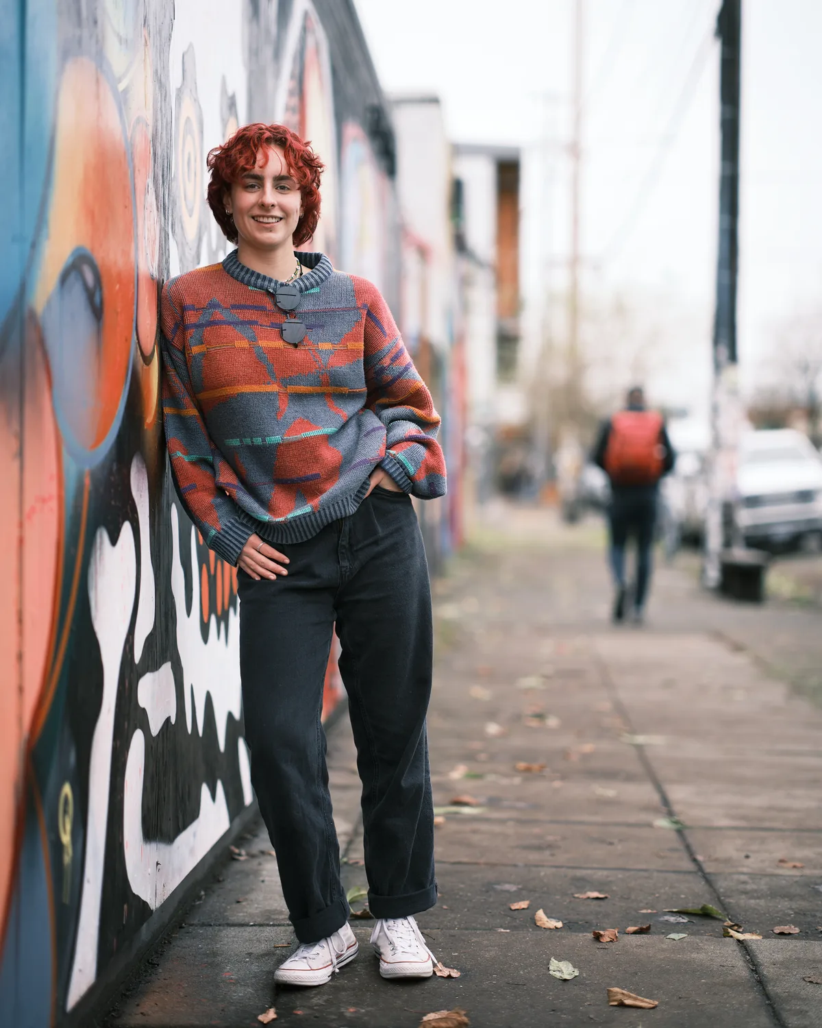 A full-length senior portrait of Annika Molina leaning against a colorful street mural on a Portland sidewalk. She has short curly red hair and wears an oversized patterned sweater in orange, grey, and teal, dark wide-leg jeans cuffed at the ankle, white Converse sneakers, and a pendant necklace. She smiles warmly at the camera with one hand on her hip. A pedestrian with a red backpack walks in the blurred background along the tree-lined street.