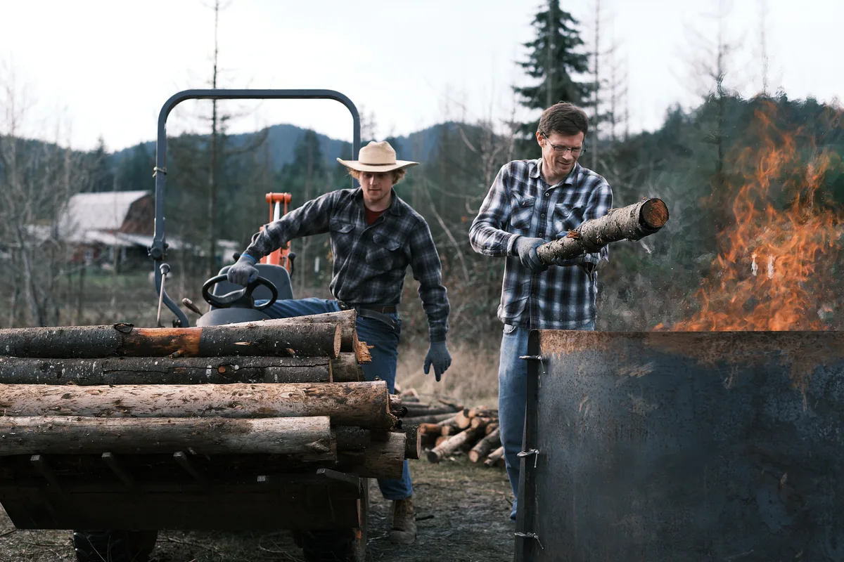 Two farmers in matching plaid flannel shirts and work gloves make biochar on a rural property. The man on the right feeds a log into a large metal burn barrel with open flames, while the younger man on the left, wearing a straw cowboy hat, tends to a pile of logs on a trailer. A chainsaw rests on the trailer nearby. A barn, bare-limbed trees, and forested hills are visible in the background under an overcast sky.