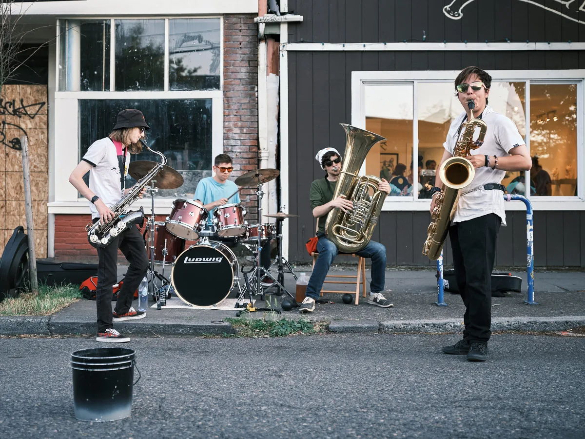 Four young musicians perform as a street band on Alberta Street during Last Thursday in Portland, Oregon. From left to right, a saxophonist in a bucket hat and white t-shirt plays tenor saxophone, a drummer in a light blue t-shirt plays a red Ludwig kit, a tuba player in a green shirt and sunglasses sits on a stool, and a baritone saxophonist in a white shirt and sunglasses plays in the foreground. A black tip bucket sits on the street in front of them. Storefronts and a gallery with people visible inside form the backdrop.