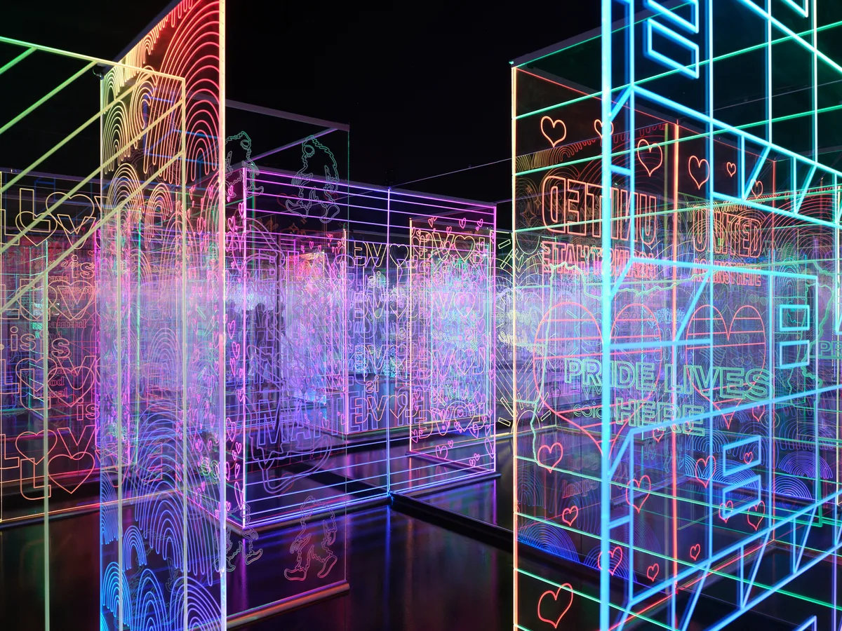 A vibrant interactive art installation featuring glowing neon geometric shapes, wireframe structures, and heart symbols in blues, pinks, and greens displayed in a dark gallery space. The luminous digital art creates an immersive environment with overlapping transparent panels and geometric forms.