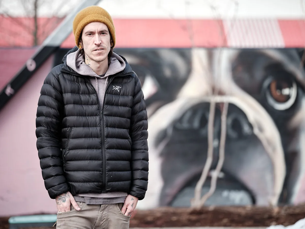 A portrait of muralist Kyle Danley, also known as Wetiko, standing in front of one of his large-scale murals depicting a close-up of a French Bulldog's face on a pink building. He wears a black Arc'teryx puffer jacket, a grey hoodie, khaki jeans, and a mustard yellow beanie. Neck and hand tattoos are visible. He looks directly into the camera with a calm, serious expression, hands in his pockets.