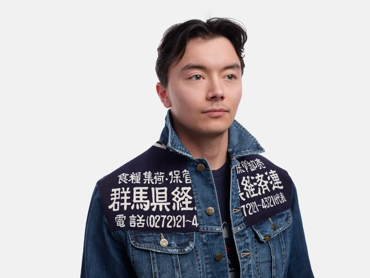 A young Asian man gazes directly at the camera with a composed, contemplative expression against a pristine white backdrop. He wears a distinctive denim jacket adorned with white Japanese characters and telephone numbers embroidered across the chest and sleeve, suggesting vintage workwear or business advertising. The even studio lighting creates soft shadows that define his features while maintaining the jacket's rich blue texture and intricate stitching details.