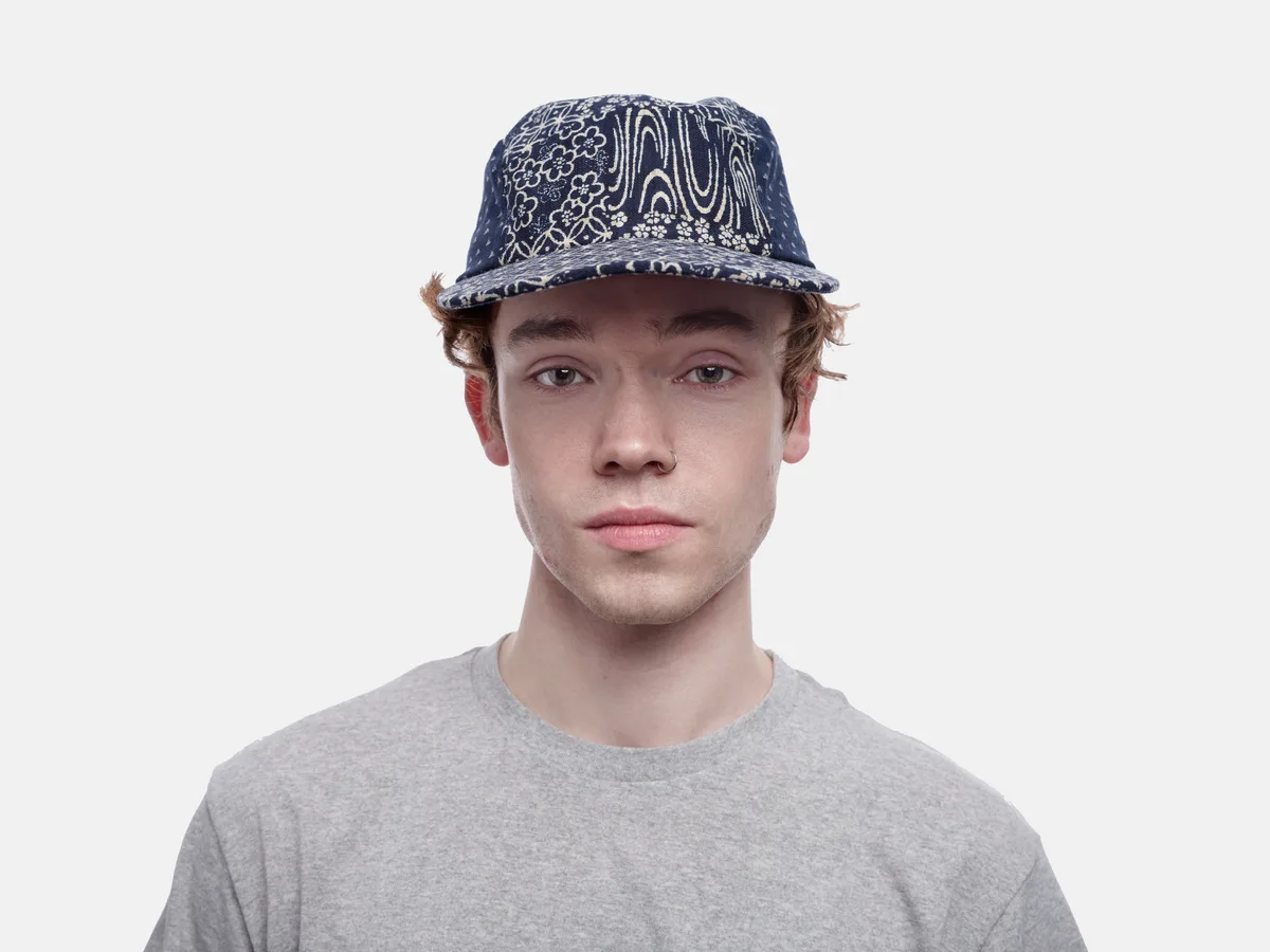 A young man with tousled blonde hair gazes directly at the camera with an earnest expression, wearing a navy blue paisley-patterned baseball cap and heather gray t-shirt. The portrait is captured against a clean white backdrop with even, diffused lighting that creates a soft, contemporary mood. His relaxed posture and steady gaze suggest casual confidence, while the intricate paisley design on his cap adds visual texture and cultural reference to the otherwise minimalist composition.