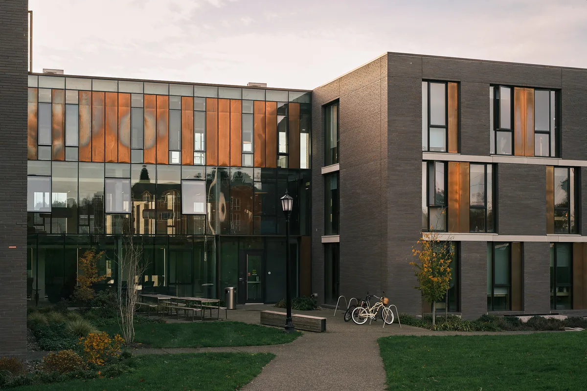 The exterior of Trillium Residence Hall at Reed College in Portland, Oregon, photographed at dusk. The modern two-story building features a striking facade of dark charcoal brick and floor-to-ceiling glass curtain walls with warm copper-toned vertical cladding that reflects the last light of the day. Older campus buildings are reflected in the large glass panels. A classic black lamppost stands at the center of a paved courtyard pathway, with two bicycles locked to a rack nearby. Outdoor benches, young autumn trees, and a well-maintained lawn complete the quiet campus scene under a softly clouded sky.