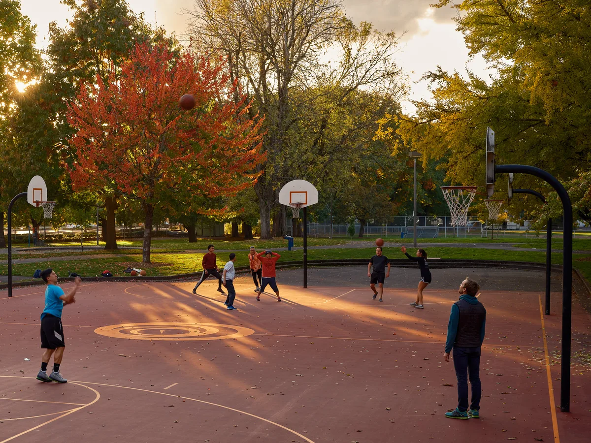 A group of people play pickup basketball on an outdoor court at Powell Park in Portland, Oregon, bathed in warm golden evening light. A basketball is suspended mid-air above the court. Three hoops are visible across the wide red-surfaced court, with fallen autumn leaves scattered across it. Vibrant fall foliage in shades of red, orange, and yellow fills the background, with the setting sun breaking through the trees on the left.