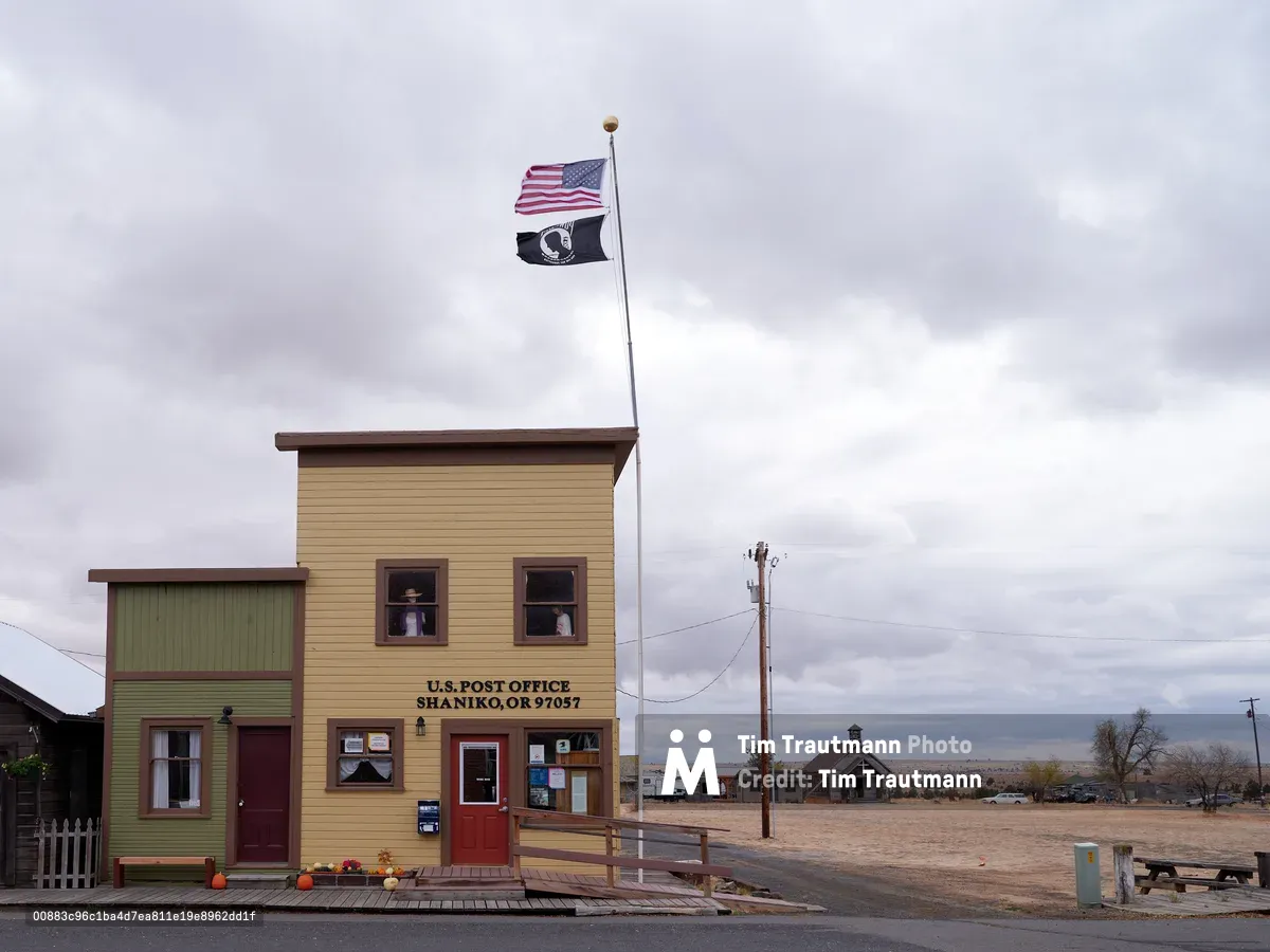 The weathered yellow clapboard post office of Shaniko, Oregon stands resolute beneath a blanket of pewter clouds, its American and POW-MIA flags snapping in the high desert wind. The modest frontier building, with its sage green trim and red door, anchors a desolate stretch of the Shaniko Historic District where time seems suspended between past and present. Autumn pumpkins dot the wooden boardwalk while telephone poles stretch into the vast Oregon plateau, their lines cutting through the moody overcast sky that threatens rain over this ghost town remnant.