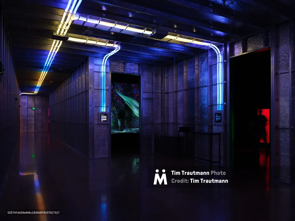 A dramatic interior hallway at Hopscotch in Portland, Oregon features industrial architecture with exposed brick walls, neon blue and purple lighting strips running along the ceiling and walls, creating a cyberpunk atmosphere. The corridor leads to what appears to be an entertainment area with glowing screens and digital displays visible in the distance.