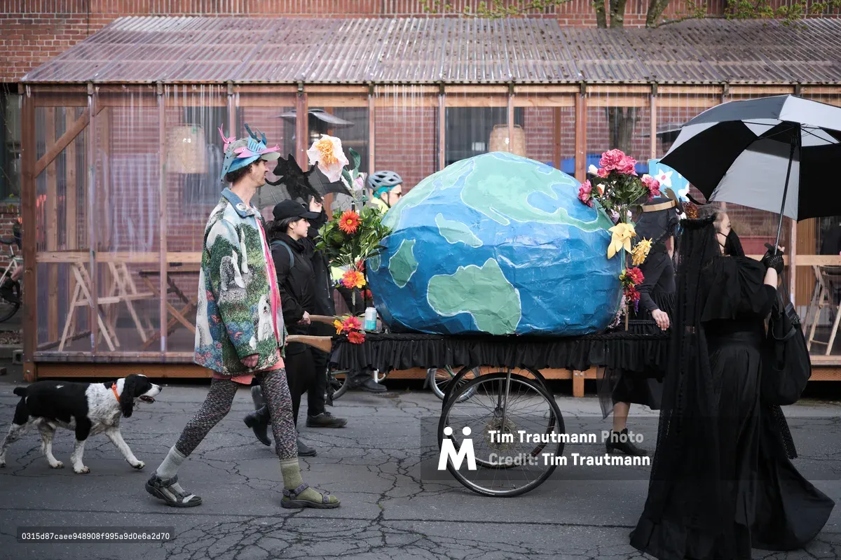A group of mourners dressed in black carry a large blue and green Earth-shaped coffin on a wheeled cart through the streets of Portland, Oregon, accompanied by flowers and an umbrella in what appears to be an environmental protest or artistic funeral procession.