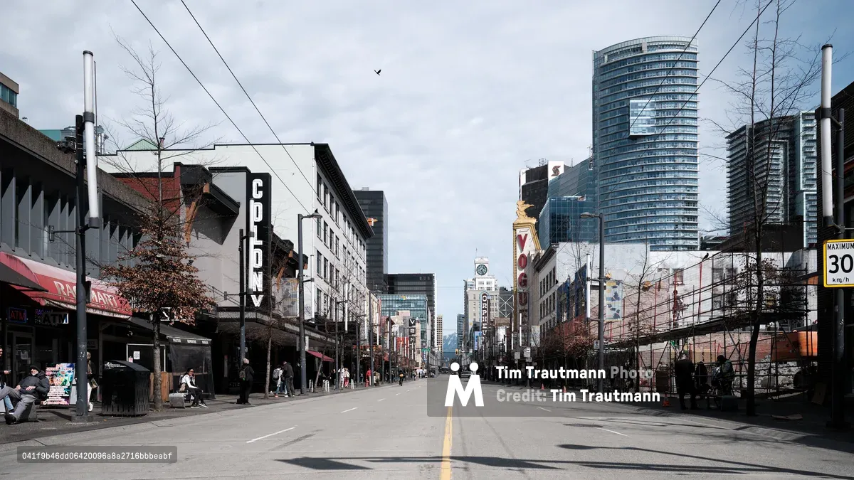 A deserted asphalt boulevard stretches toward Vancouver's downtown core, flanked by the eclectic mix of heritage storefronts and soaring glass towers that define the city's evolving skyline. The Colony Theatre's bold vertical signage punctuates the left side of the frame, while modern residential high-rises pierce the overcast sky in the distance. Sparse pedestrians and the conspicuous absence of traffic create an unusual quietude along this typically bustling commercial artery, with power lines creating geometric patterns overhead against the muted gray clouds.