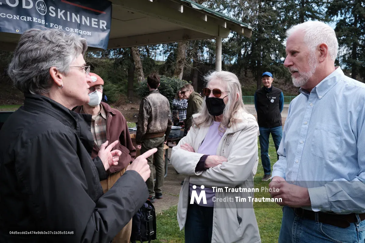 A group of people gathered outdoors at a political campaign event in Oregon City, Oregon, with a campaign banner visible in the background. The scene shows several individuals engaged in conversation, some wearing masks, in what appears to be a park setting with trees and grass.