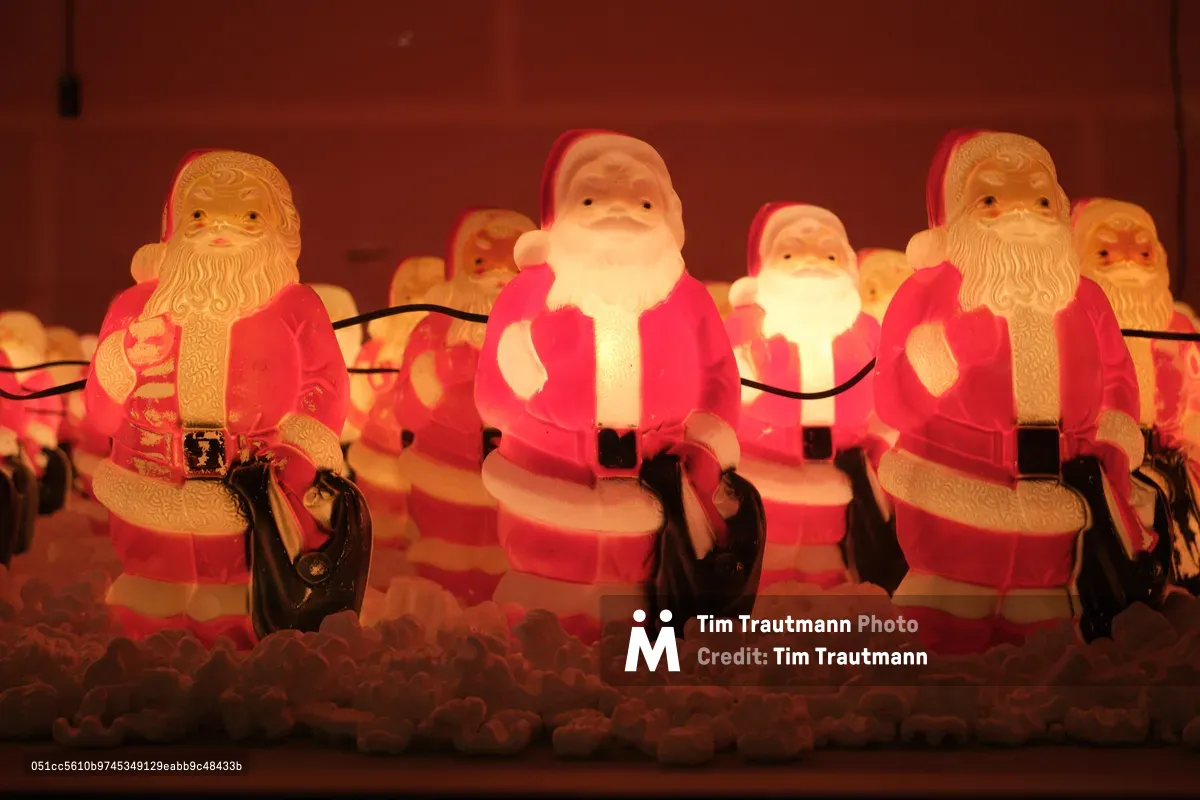 A mesmerizing battalion of identical illuminated Santa Claus figures stands sentinel in Portland's Concordia neighborhood, their warm amber glow piercing the burgundy winter darkness. Each plastic saint clutches a black lantern, creating a rhythmic pattern of jolly faces that stretches into the atmospheric haze. The figures emit an ethereal radiance against the moody evening sky, transforming Northeast Killingsworth Street into a surreal Christmas dreamscape where mass-produced holiday cheer takes on an almost mystical quality.