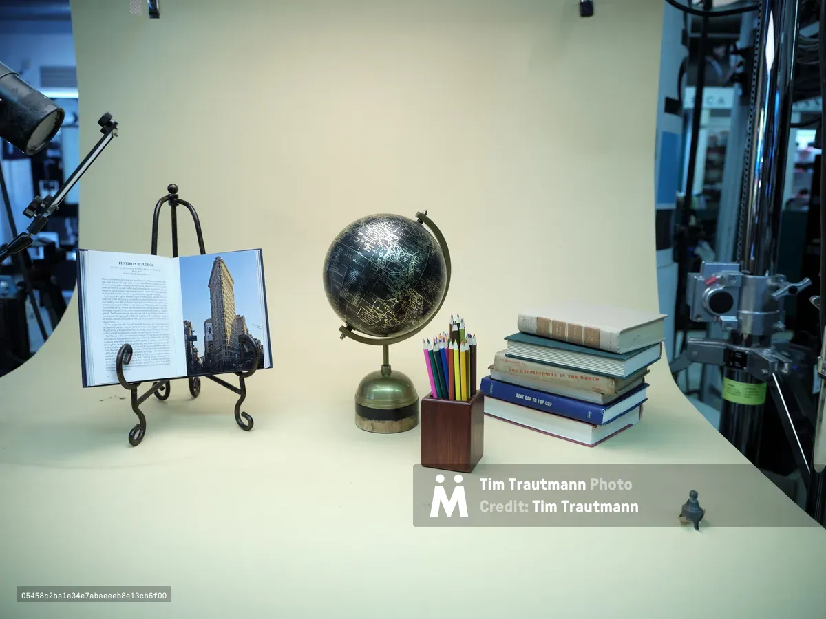 A meticulously arranged still life captures the essence of intellectual pursuit on a pristine white surface within a professional photography studio. The composition centers around a dark celestial globe with golden meridian rings, flanked by a stack of weathered books, an open architectural volume displaying the iconic Flatiron Building, and a wooden pencil holder bristling with colorful writing instruments. Studio lighting creates subtle shadows and highlights against the neutral backdrop, while professional equipment frames the edges, revealing the careful orchestration behind this contemplative tableau.