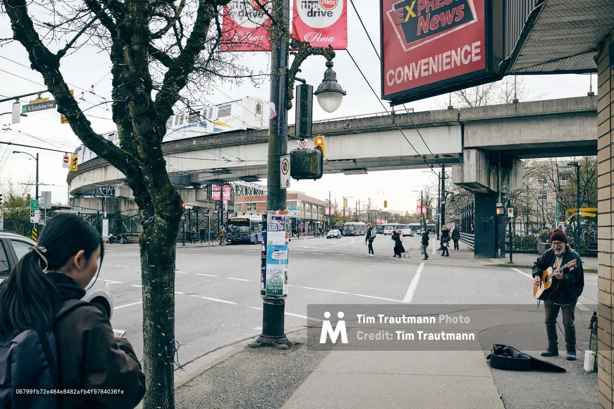 A street musician with an acoustic guitar performs beneath the weathered concrete spans of the North Grandview Highway overpass in Vancouver's Grandview-Woodland neighborhood. The moody overcast sky casts soft, diffused light over the urban intersection, where pedestrians cross the wide street and a hooded figure in the foreground observes the scene. Red convenience store signage and bare winter trees frame this intimate moment of street performance against the backdrop of Vancouver's transit infrastructure.