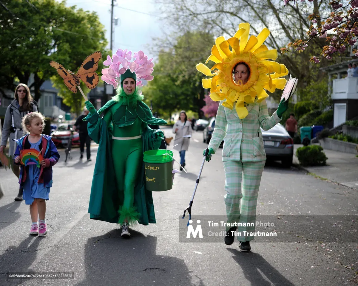 Two people in elaborate flower costumes walk down a residential street in Portland's Central Eastside district during what appears to be an environmental awareness parade or community event. One person wears a green outfit with a pink flower headpiece and carries a bucket labeled 'Kitchen Planet', while the other is dressed in a yellow sunflower costume with matching plaid suit.