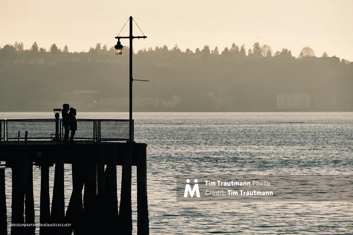 Two figures stand in intimate silhouette against the amber-washed waters of Elliott Bay from Seattle's Pier 70, their forms darkened against the ethereal golden hour light. A vintage lamp post rises beside them like a sentinel, while the distant shoreline dissolves into layers of atmospheric haze. The weathered wooden pier pilings create strong vertical lines that anchor the composition, as gentle ripples catch the dying light across the bay's surface.