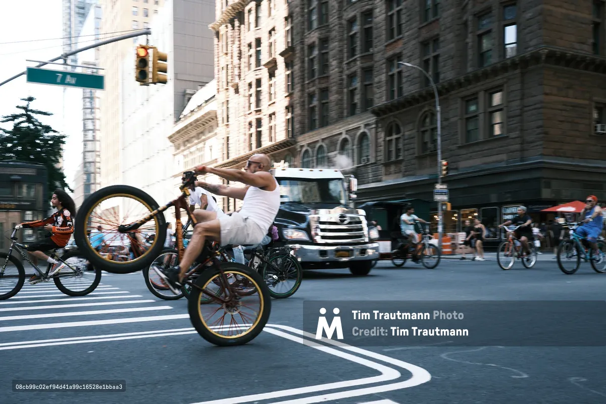 A shirtless cyclist in white shorts performs a dramatic wheelie on his custom bicycle, commanding the center of 7th Avenue in Midtown Manhattan. The golden hour light bathes the historic architecture and creates dynamic shadows across the bustling intersection, while traffic and pedestrians blur into the urban rhythm. The scene captures the raw energy and improvisational spirit of New York street culture against the towering backdrop of Manhattan's iconic skyline.
