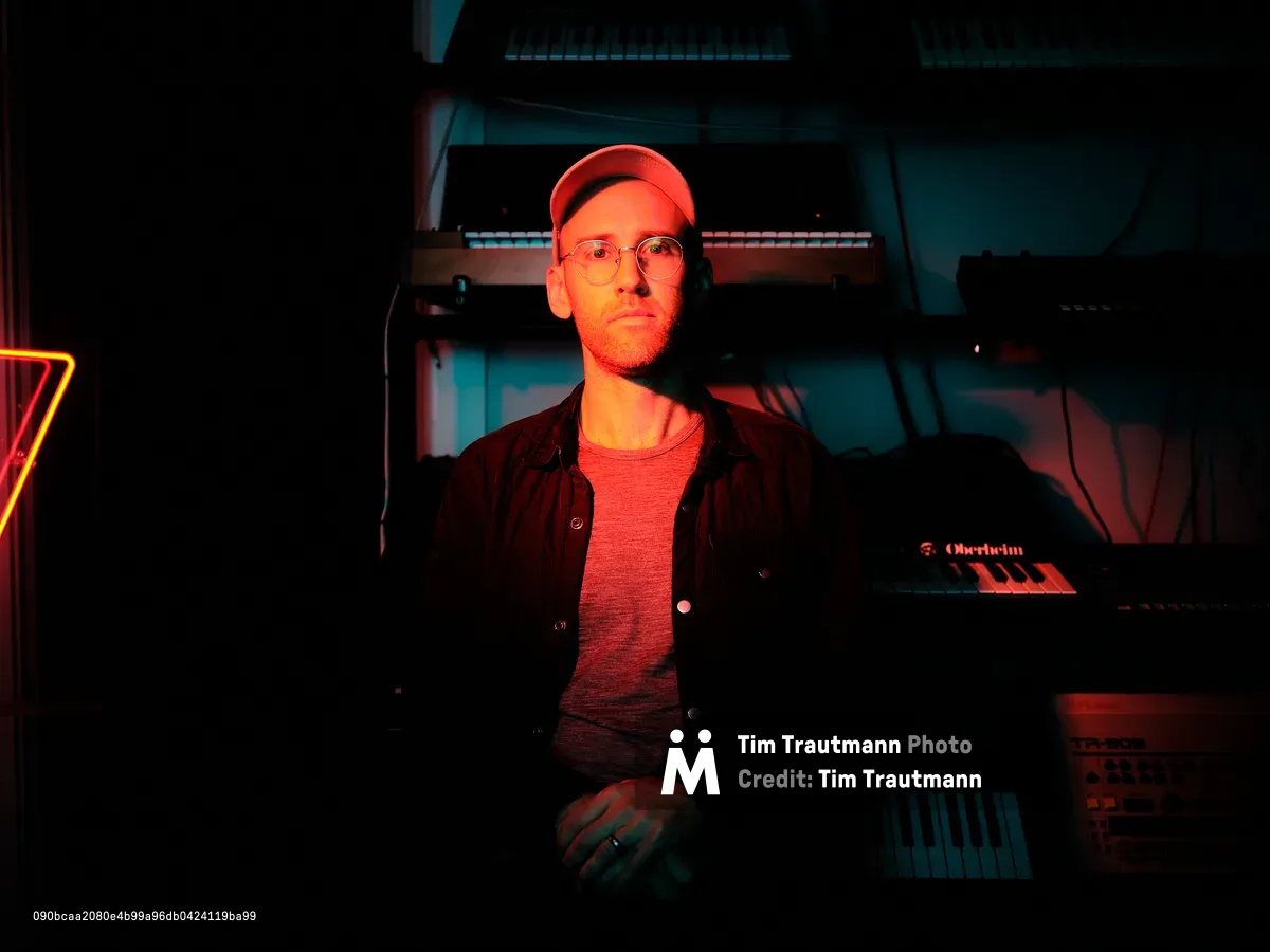 A portrait of DJ and event producer Peter Marks seated in a darkened music studio, lit dramatically by warm red light. He wears round glasses, a baseball cap, a grey t-shirt, and an open dark shirt. An Oberheim synthesizer and a Roland TR-909 drum machine are visible in the foreground, with additional keyboards and equipment stacked behind him. A red neon triangle glows on the left side of the frame.