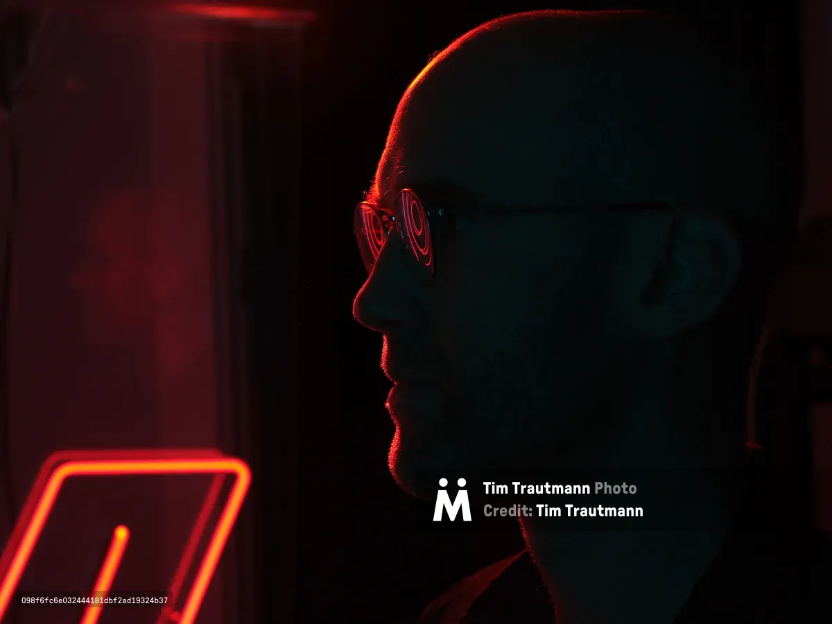 A silhouetted profile of a person wearing glasses illuminated by dramatic red and orange neon lighting in a dark urban setting. The atmospheric lighting creates a moody, cyberpunk-style scene with glowing neon signs visible in the background.