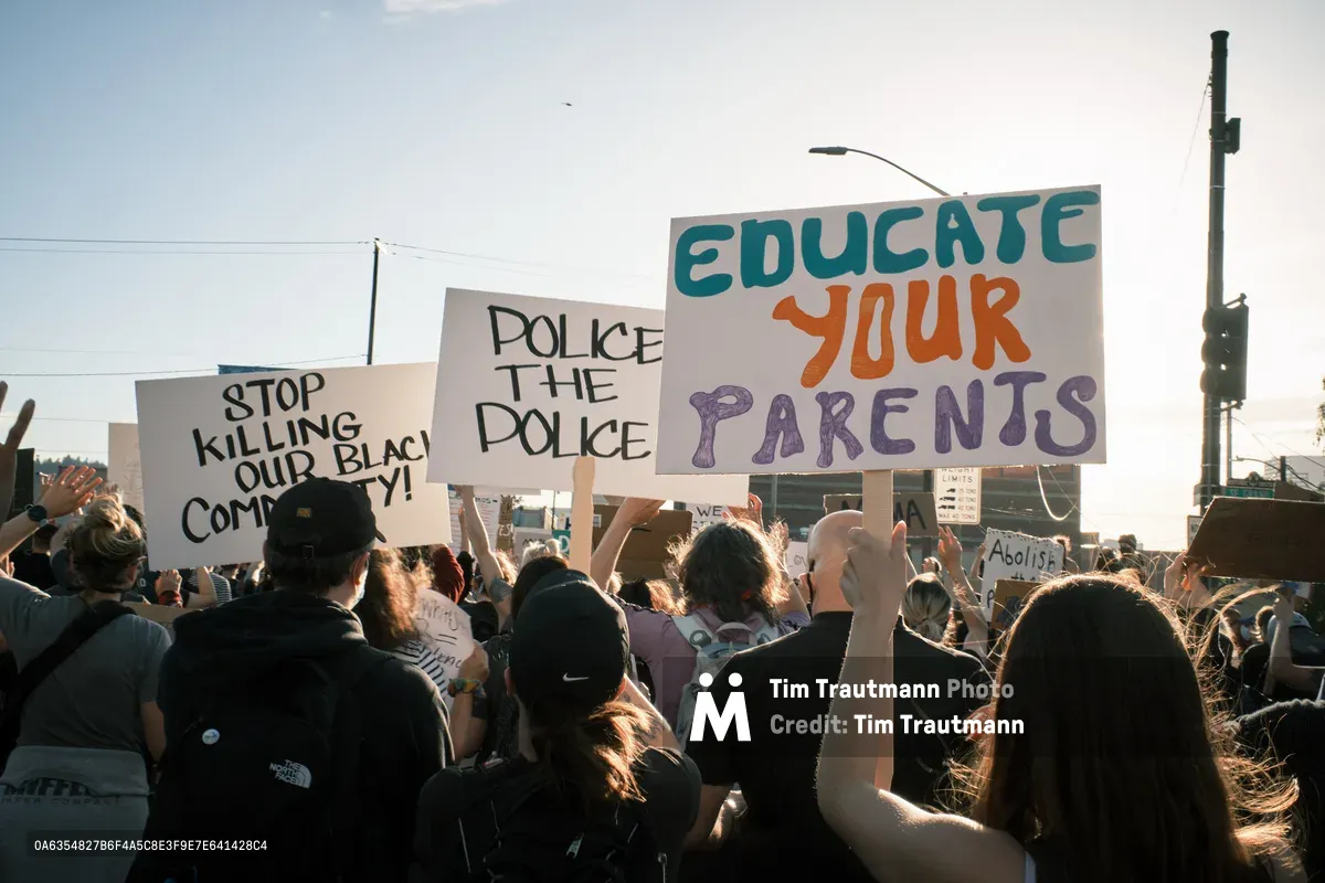 A dense crowd of protesters moves purposefully across Portland's Morrison Bridge, their handmade signs catching the golden hour light against a pale sky. The demonstrators hold messages including "STOP KILLING OUR BLACK COMMUNITY," "POLICE THE POLICE," and "EDUCATE YOUR PARENTS," their colorful lettering standing out against white poster board. The late afternoon sun backlights hair and shoulders, creating an atmosphere of determined solidarity as the group advances toward downtown Portland. Power lines and urban infrastructure frame the scene, emphasizing the grassroots nature of this civil rights demonstration.