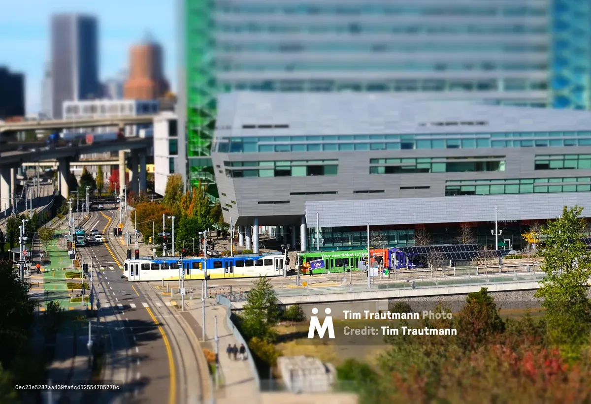 A tilt-shift photograph of Portland's South Waterfront district, giving the scene a miniature toy-like appearance. A TriMet MAX light rail train in blue and yellow livery runs along the street-level tracks in front of the OHSU Collaborative Life Sciences Building, a large modern structure clad in grey panels and green glass. A green-wrapped advertising train and a purple bus are visible at the adjacent transit stop. An elevated freeway interchange curves through the background, with the blurred Portland downtown skyline and autumn foliage visible in the distance under a clear blue sky.