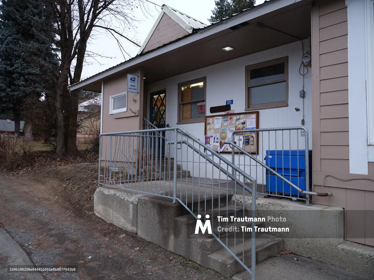 A modest single-story post office building stands quietly in Ronald, Washington, its beige siding and utilitarian design embodying the practical architecture of small American towns. Metal railings guide visitors up concrete steps to the entrance, where a bulletin board heavy with community notices speaks to the building's role as a local gathering point. The overcast Pacific Northwest sky and bare winter trees frame this essential civic outpost, while the building's simple geometry and muted palette reflect the unpretentious character of rural postal service.