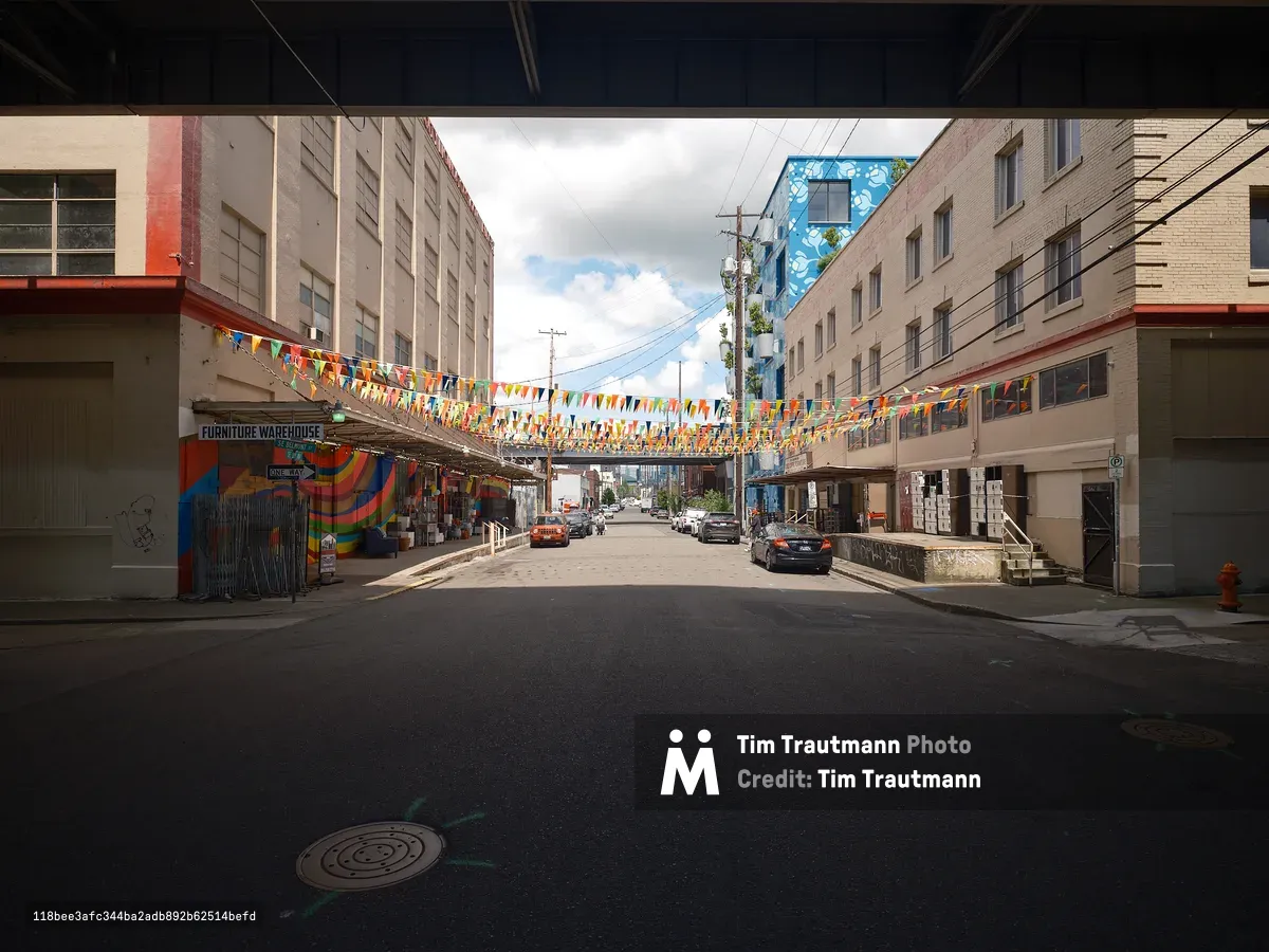 A street-level view looking down a narrow urban block beneath a freeway overpass in Portland, Oregon, outside City Liquidators furniture warehouse. Colorful rainbow bunting flags are strung across the street between the buildings on both sides. A vibrant rainbow-colored mural decorates the left side of the building, with a "Furniture Warehouse" sign visible above it. In the background, a blue building covered in a large floral mural rises above the streetscape. A few parked and moving cars occupy the quiet cobblestone street, and a partly cloudy sky is visible between the buildings ahead.