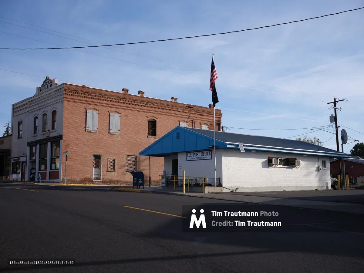 The modest U.S. Post Office in Dufur, Oregon sits beneath a sweeping blue sky streaked with wispy clouds, its bright blue roof a striking contrast against the weathered brick buildings of this small prairie town. Power lines trace geometric patterns overhead while an American flag hangs limp in the still air, creating a quintessentially American small-town tableau. The empty main street stretches into shadow, capturing the quiet dignity of rural postal service in the Columbia River Gorge region.