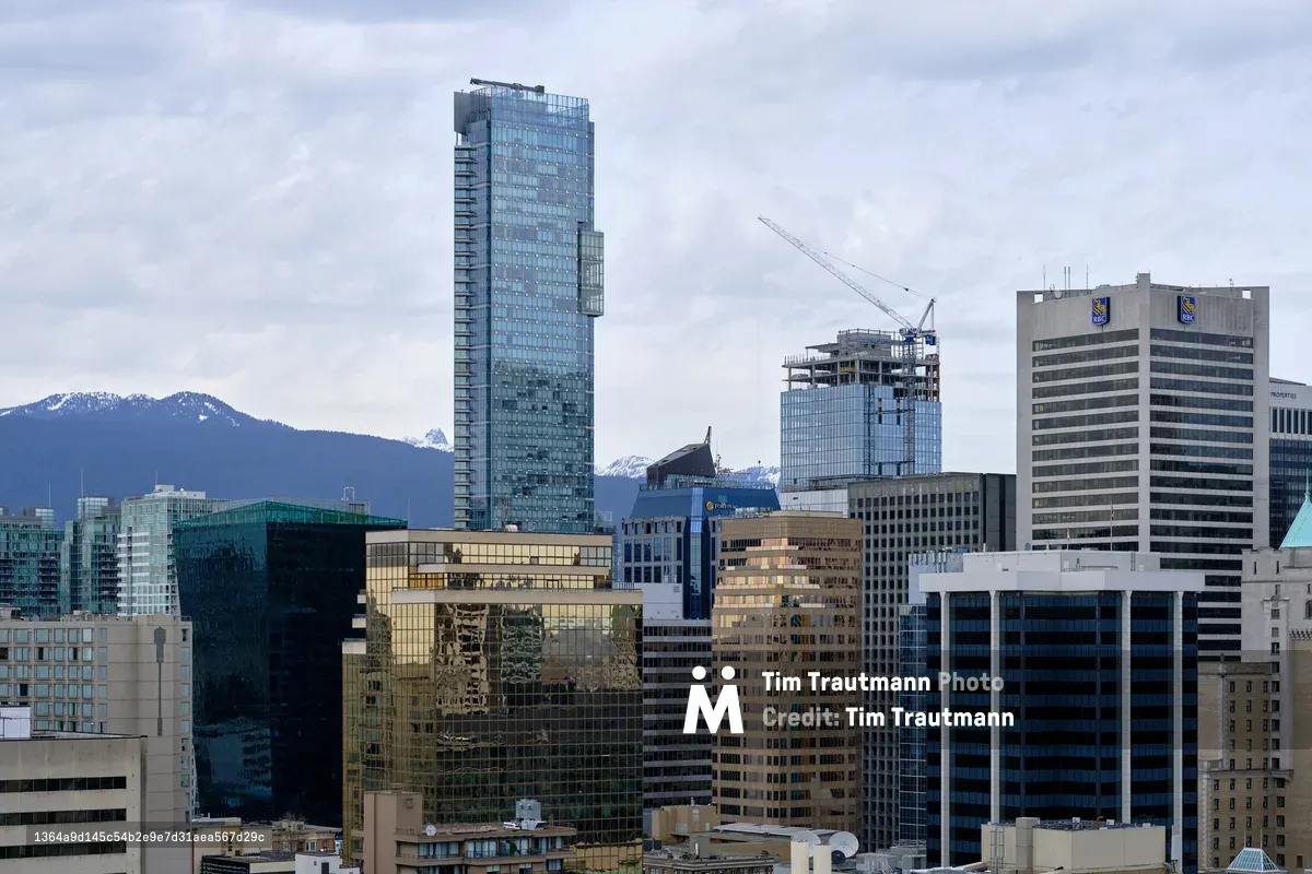 The gleaming downtown core of Vancouver rises in sharp architectural contrast against the snow-dusted North Shore Mountains. A commanding blue glass tower dominates the skyline while construction cranes hint at the city's relentless growth. The overcast Pacific Northwest sky creates a muted palette that emphasizes the interplay between urban ambition and natural grandeur, with the coastal range providing a dramatic backdrop to this dense collection of office buildings and residential towers.