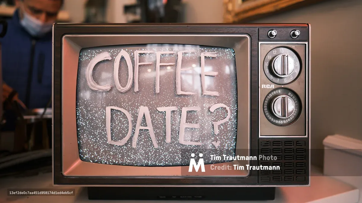 A weathered RCA television set from the 1970s displays the romantic query 'COFFEE DATE?' in elegant white lettering against a dreamy, star-speckled backdrop of soft pinks and blues. The vintage wood-grain console, complete with analog dials and speaker grille, sits in the intimate atmosphere of Edge of the Circle Books in Seattle's University District. Warm ambient lighting from the bookstore creates a nostalgic glow, while the blurred figure of a patron in navy clothing adds human presence to this whimsical intersection of analog technology and modern courtship.