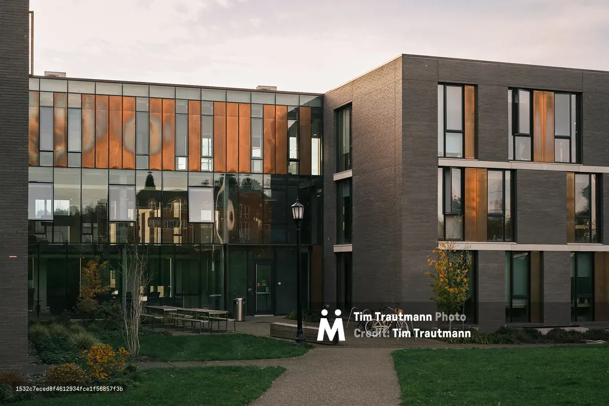 The exterior of Trillium Residence Hall at Reed College in Portland, Oregon, photographed at dusk. The modern two-story building features a striking facade of dark charcoal brick and floor-to-ceiling glass curtain walls with warm copper-toned vertical cladding that reflects the last light of the day. Older campus buildings are reflected in the large glass panels. A classic black lamppost stands at the center of a paved courtyard pathway, with two bicycles locked to a rack nearby. Outdoor benches, young autumn trees, and a well-maintained lawn complete the quiet campus scene under a softly clouded sky.