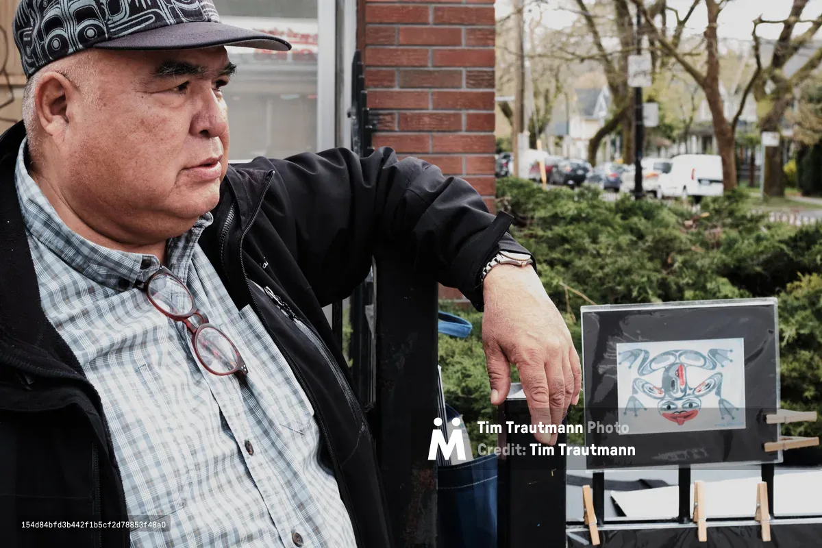 Brad Henry, a former firefighter turned traditional Tlingit artist, sits contemplatively beside his artwork on Commercial Drive in Vancouver's Grandview-Woodland neighborhood. The Indigenous artist, wearing a distinctive patterned cap and plaid shirt beneath a dark jacket, gestures toward a framed piece featuring traditional blue and red Tlingit designs. Natural daylight filters through the urban setting, highlighting the weathered brick architecture and spring foliage that frame this intimate portrait of cultural preservation and artistic dedication.